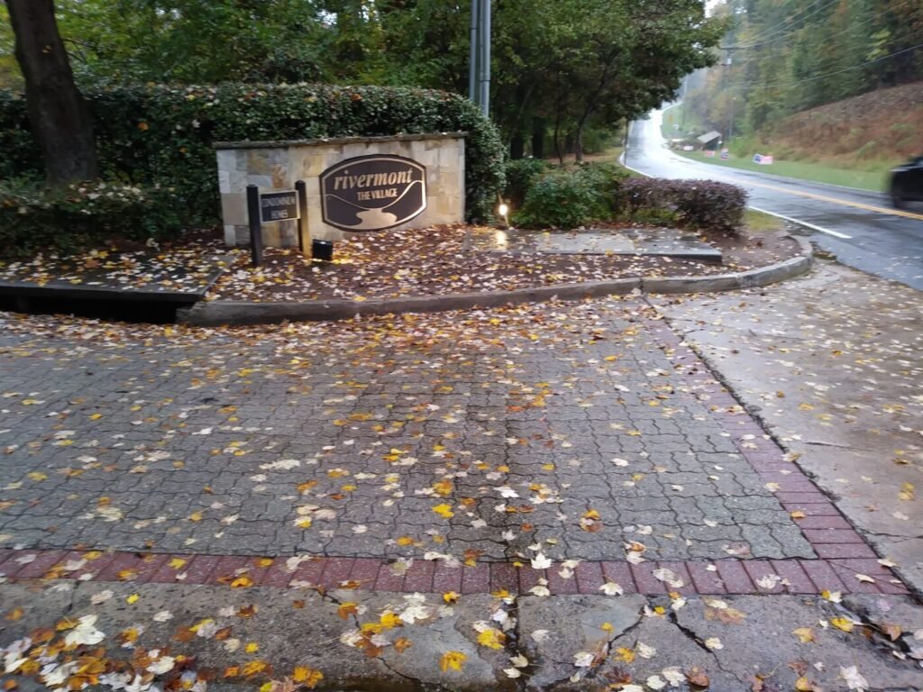 Rivermont The Village neighborhood entrance sign in Johns Creek, Georgia, along a tree-lined road with autumn leaves on the ground.