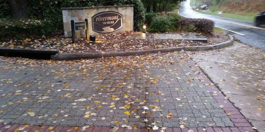 Rivermont The Village neighborhood entrance sign in Johns Creek, Georgia, along a tree-lined road with autumn leaves on the ground.