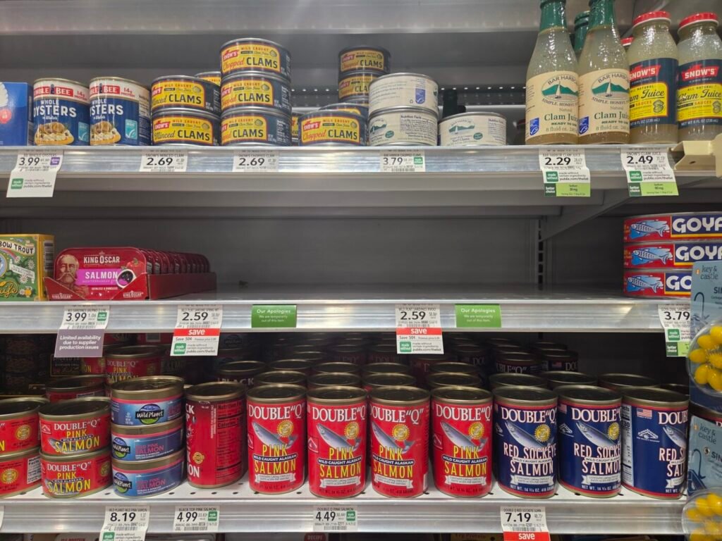 Grocery shelf with canned seafood, including clams, salmon and sardines, with some spaces between products.