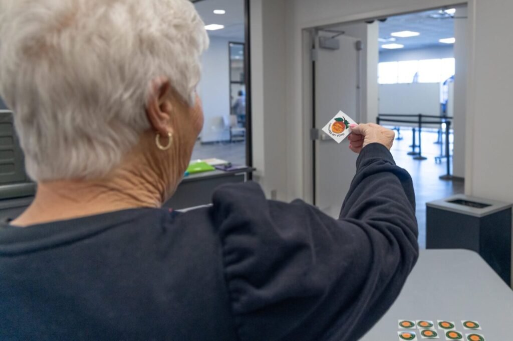 A poll worker extends a voter sticker toward an open doorway inside a polling place.