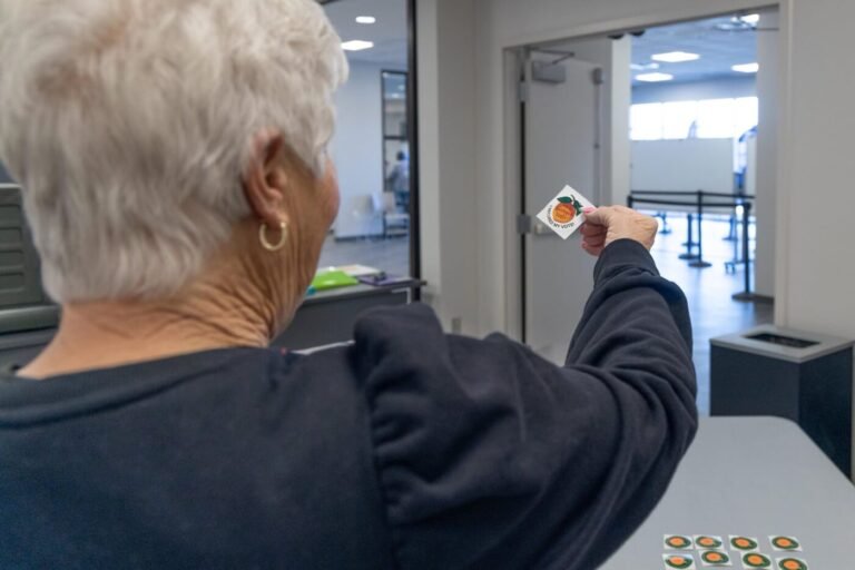 A poll worker extends a voter sticker toward an open doorway inside a polling place.