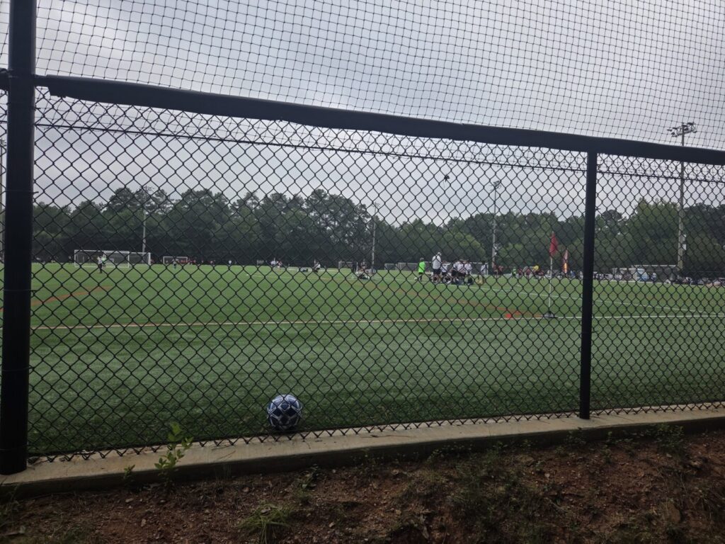 A soccer ball rests near a chain-link fence overlooking a green field with players in the distance.