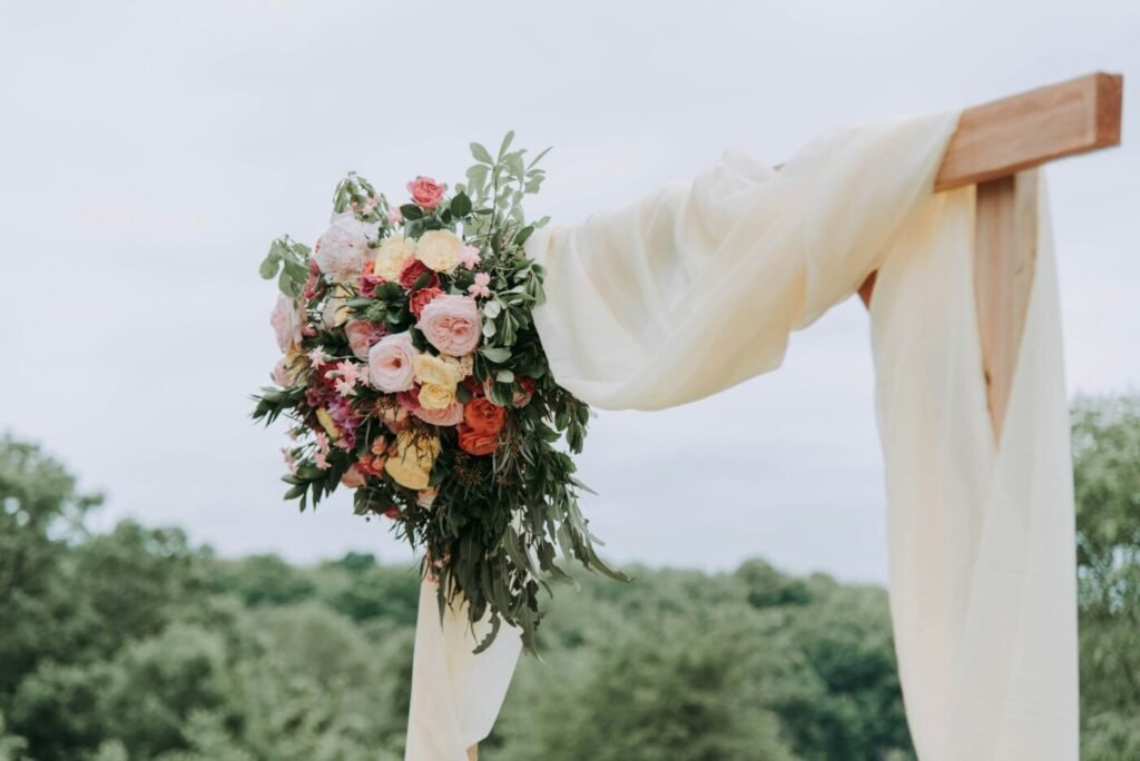 A floral arrangement and white drapery hang from a wooden wedding arch outdoors.