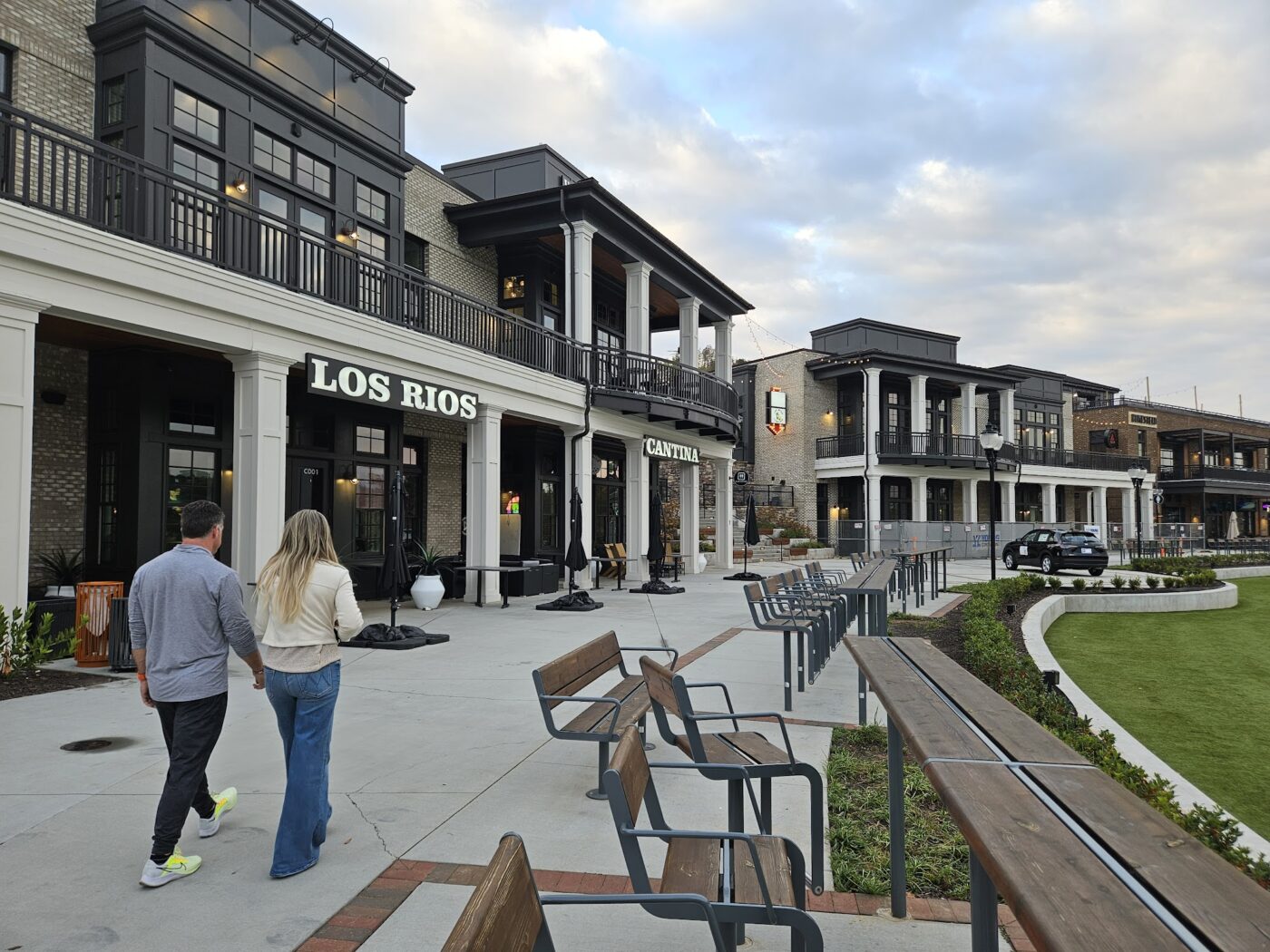Two people walk along a paved plaza beside storefronts and benches at Cumming City Center.