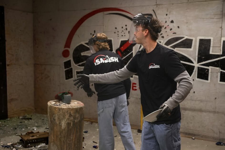 Two people wearing safety gear stand in a splatter-paint room with paint-spattered walls and a wooden stump in front of them.