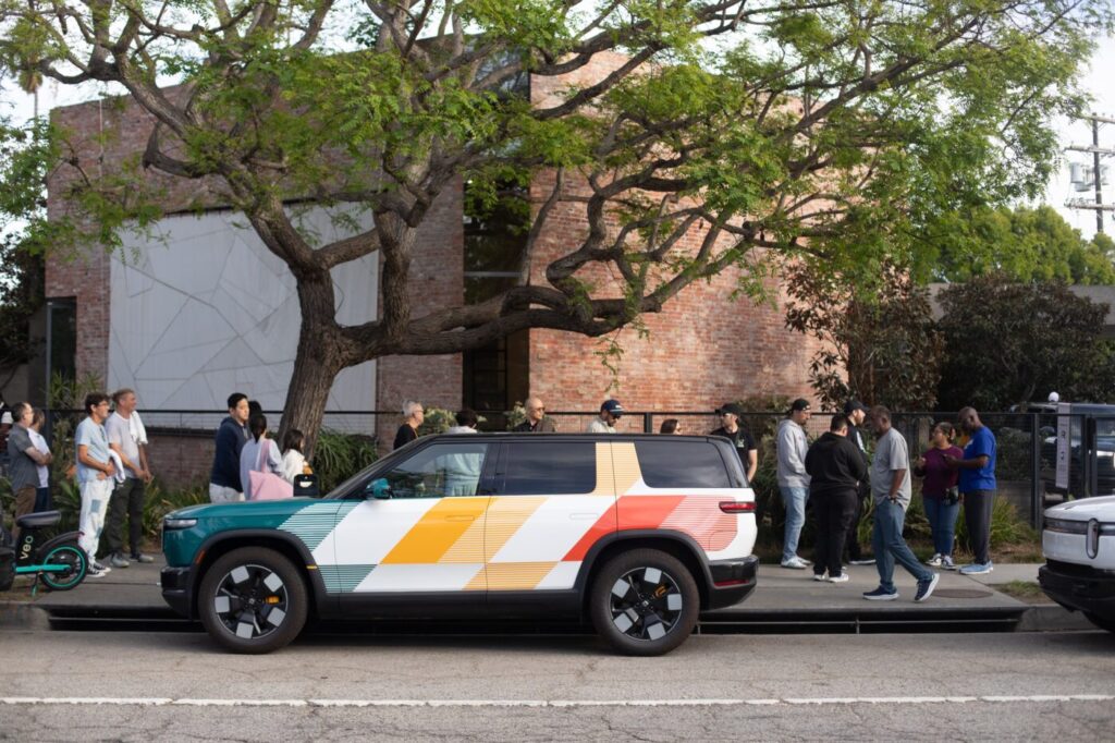 A colorful Rivian SUV parked along a curb with several people waiting on the sidewalk near a brick building and a large tree.