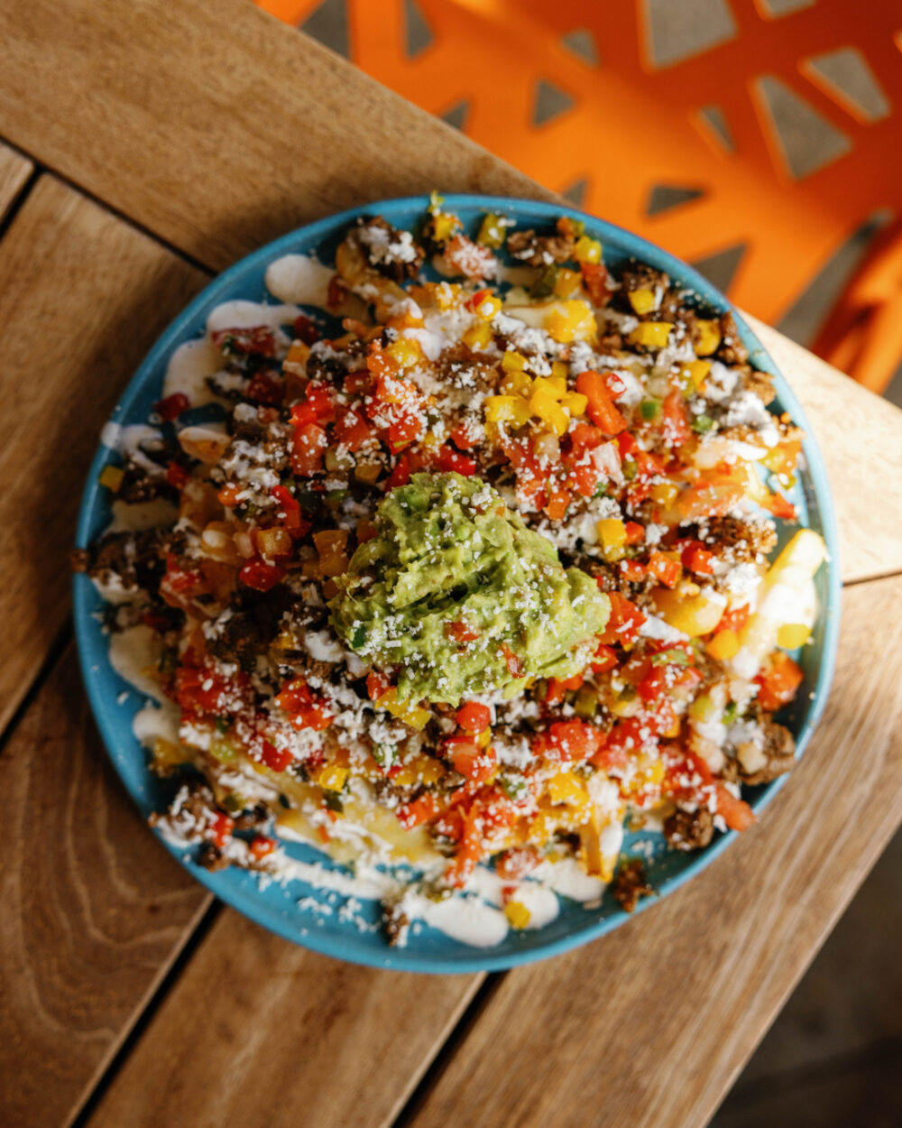 A blue plate of loaded fries topped with guacamole, diced peppers, crumbled cheese and sauce on a wooden table.