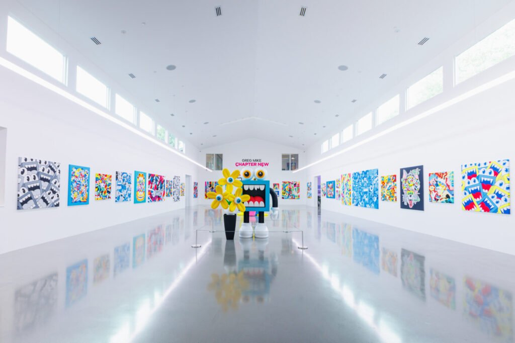 Interior view of a Greg Mike art exhibition featuring colorful contemporary paintings and large character sculptures inside a bright gallery space