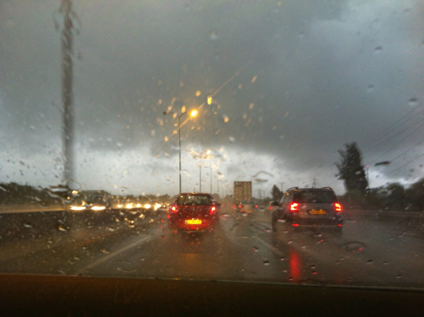 Cars drive on a wet highway in heavy rain at dusk, seen through a rain-speckled windshield.