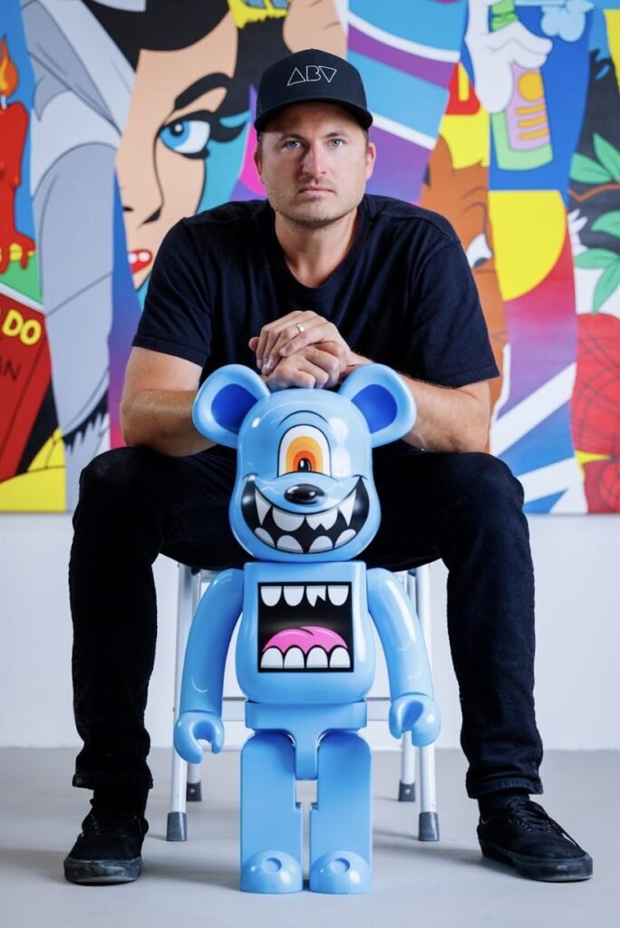Atlanta artist Greg Mike seated behind one of his signature blue character sculptures inside ABV Gallery