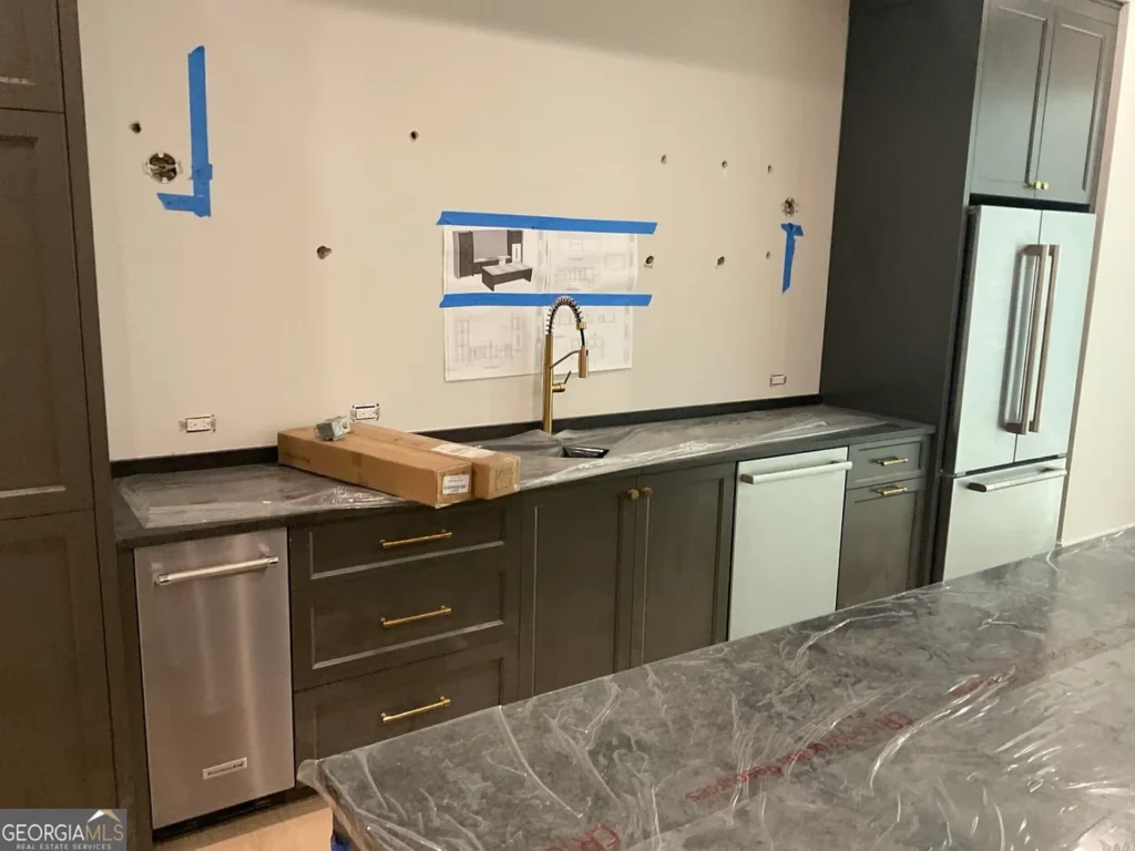 Close-up of an unfinished luxury kitchen in a Milton, Georgia estate, with gray cabinetry, marble-style counters, brass hardware, appliances and taped construction plans on the wall.