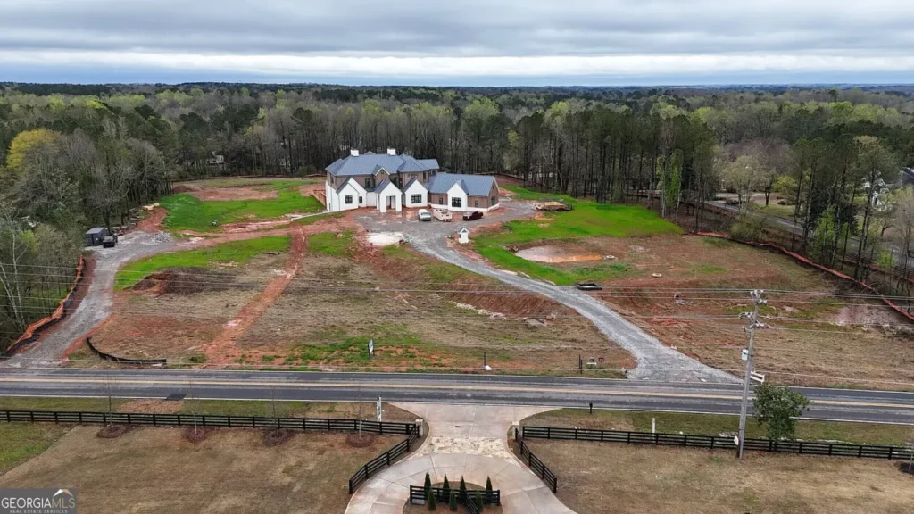 Wide aerial view of an unfinished luxury estate on a large Milton, Georgia property, with a long gravel drive, open acreage, trees and a road in the foreground.