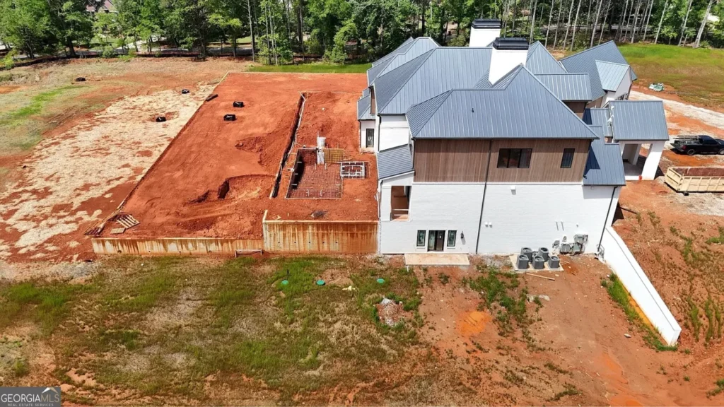 Aerial side view of a large Milton, Georgia luxury estate under construction, showing the unfinished pool area, exposed red clay, retaining wall and surrounding trees.