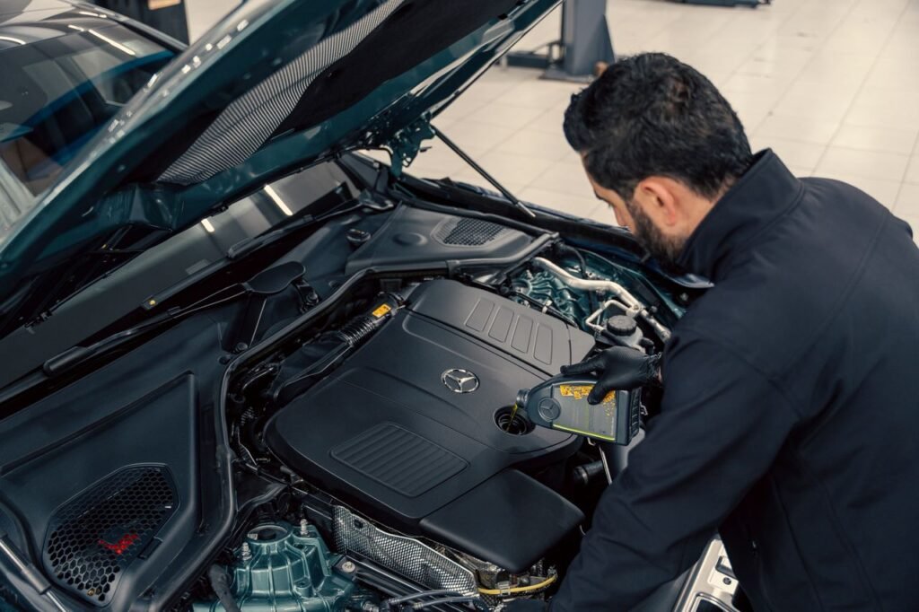 Mechanic in a dark jacket leans over an open car hood while using a handheld diagnostic tool.