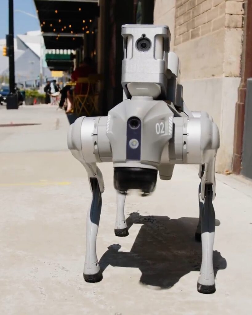 Dog-like robotic security guard walking on a city sidewalk beside a brick building.