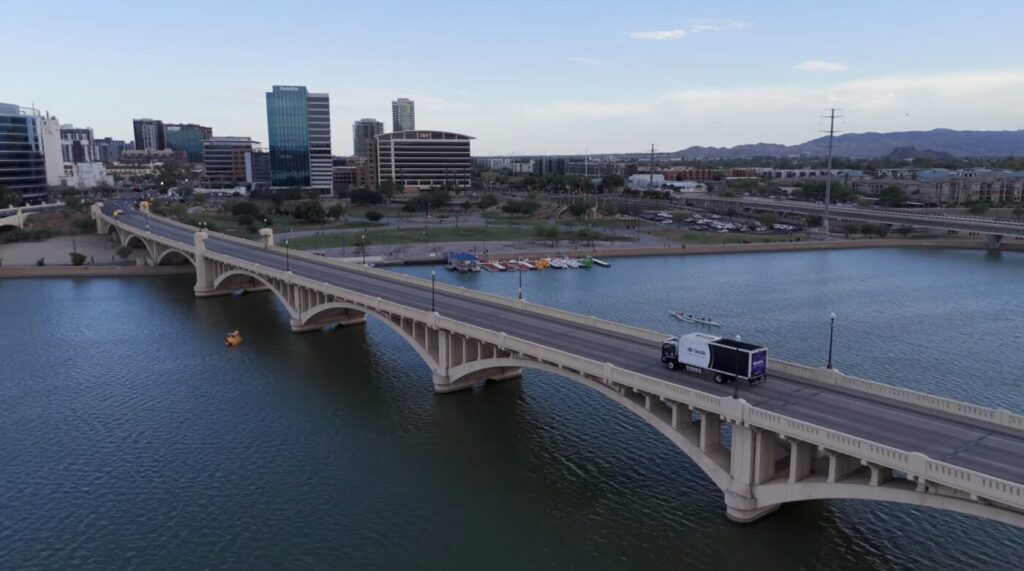 An autonomous delivery box truck drives across a bridge near an urban waterfront skyline.