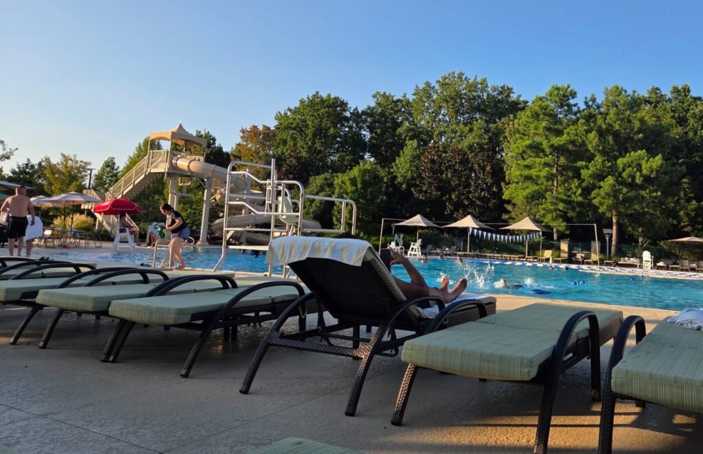 Outdoor pool area with lounge chairs, swimmers, shade umbrellas and a water slide surrounded by trees