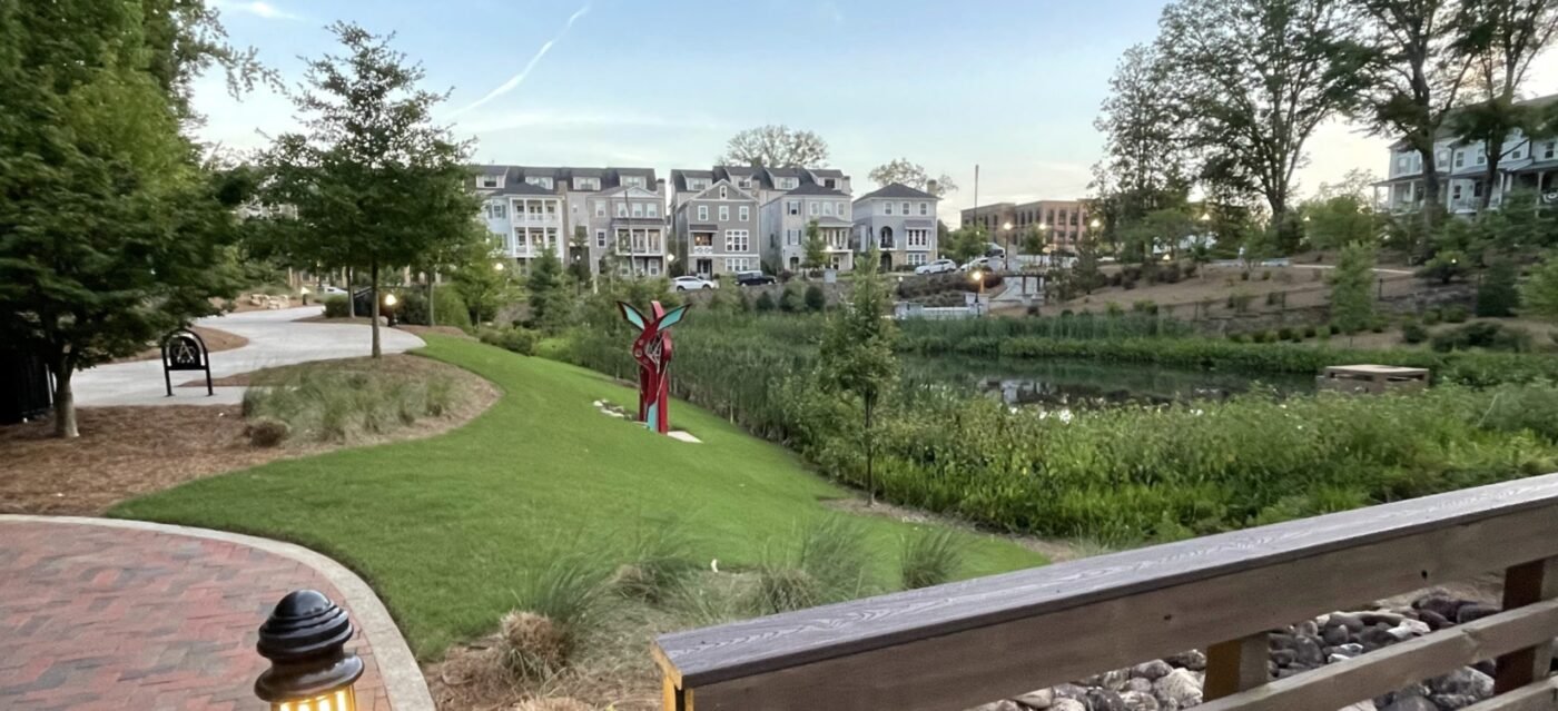Public trails and open space are central to Milton’s push to knit Deerfield into a more connected district. A greenway path curves beside a pond, with trees, a sculpture and multi-story residential buildings beyond.