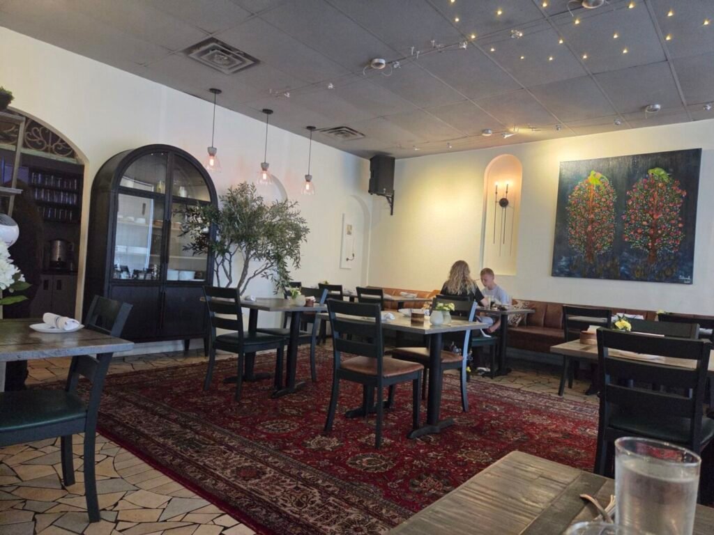 Dining room with Persian rugs, dark chairs, an olive tree, wall art and two diners at a table.