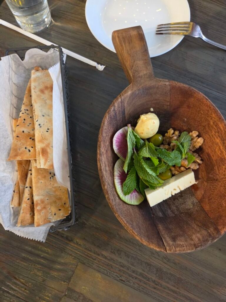 Flatbread beside a wooden bowl of feta, walnuts, olives, mint, radish and butter on a restaurant table.