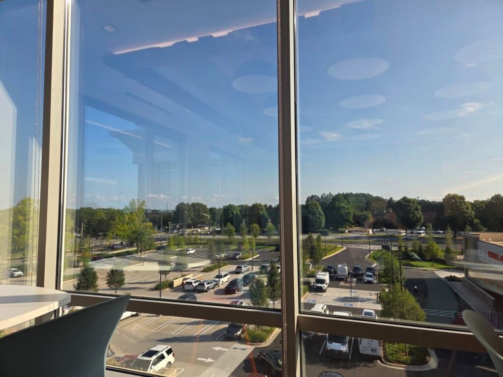 View through large office windows onto a parking area, trees and roadways under a clear sky