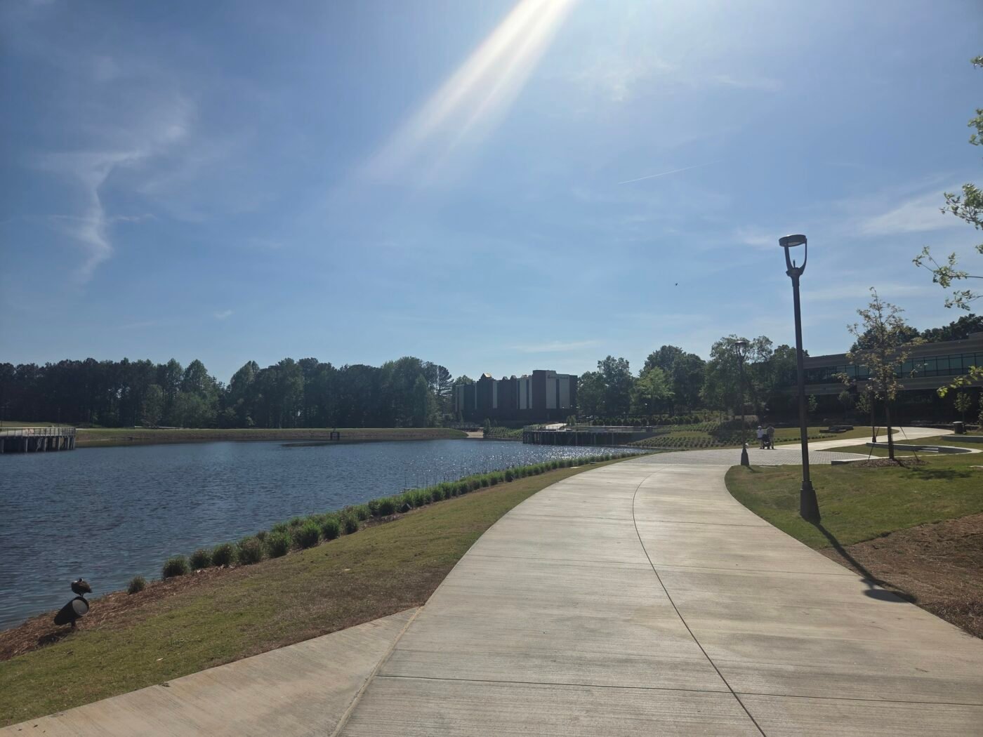 Curving paved boardwalk beside a lake with modern buildings and lampposts nearby