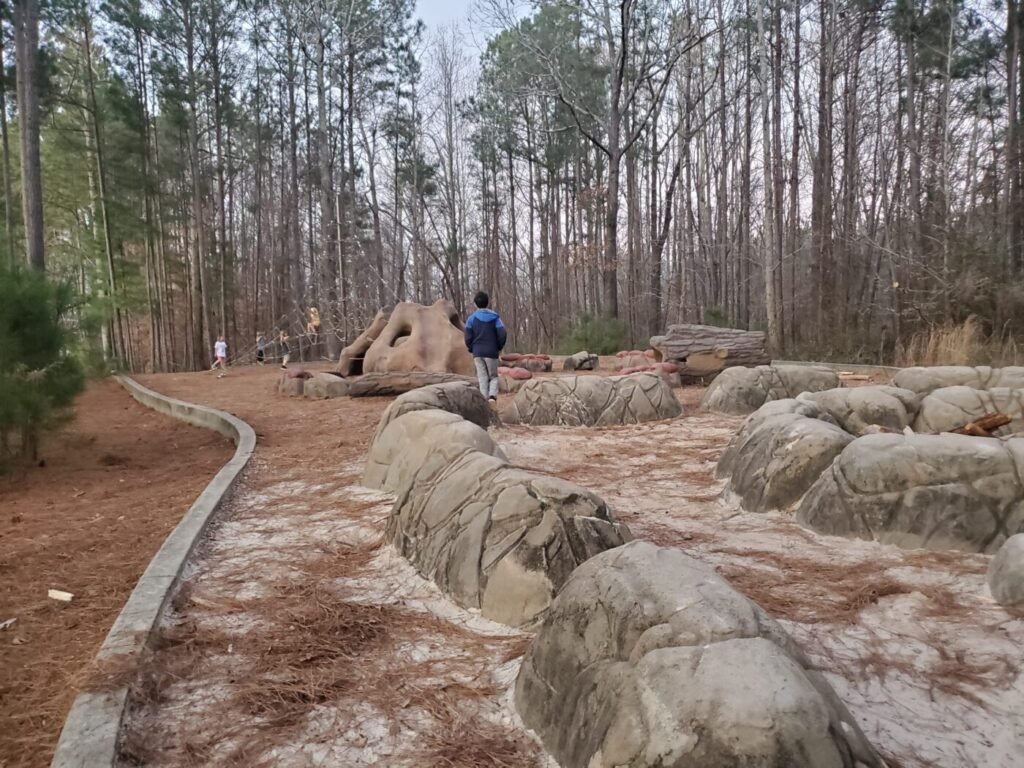 Children play among artificial rock formations in a wooded play area.