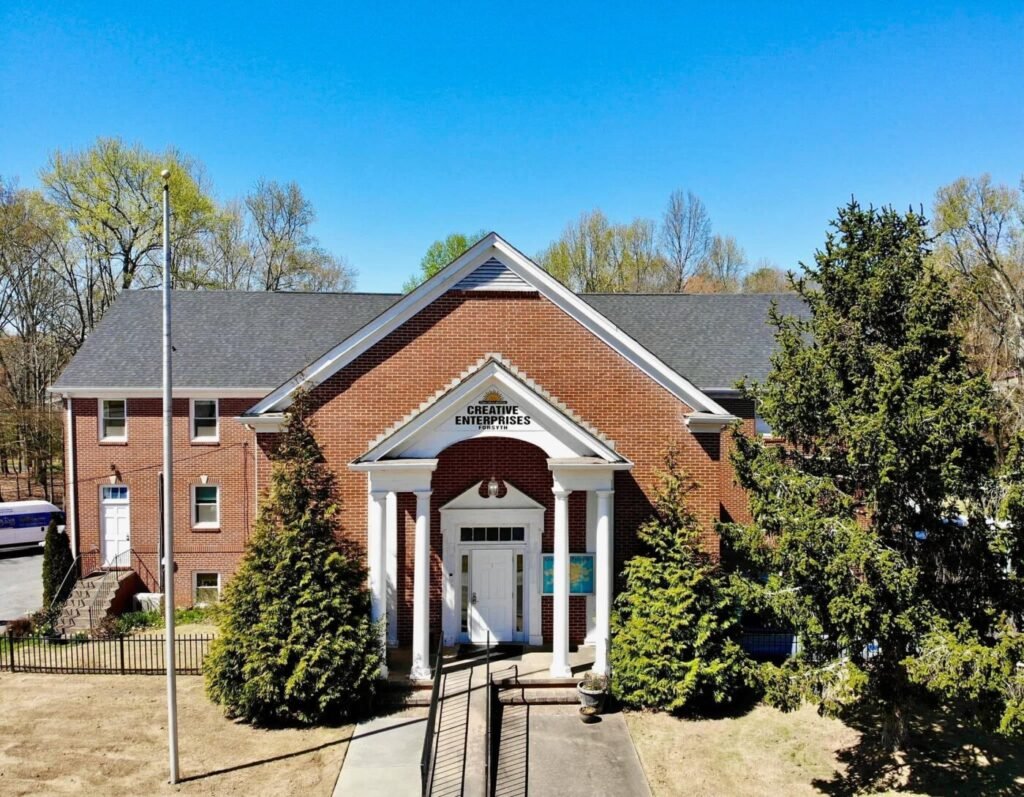 Brick building with white columns and a central entrance under a peaked roof, framed by trees and a blue sky.
