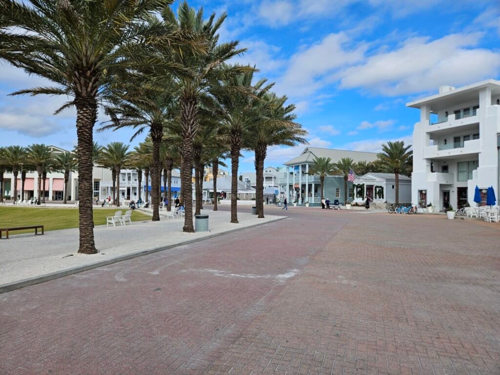 Palm trees lining a wide brick plaza with low-rise buildings and a white modern condo under a blue sky.