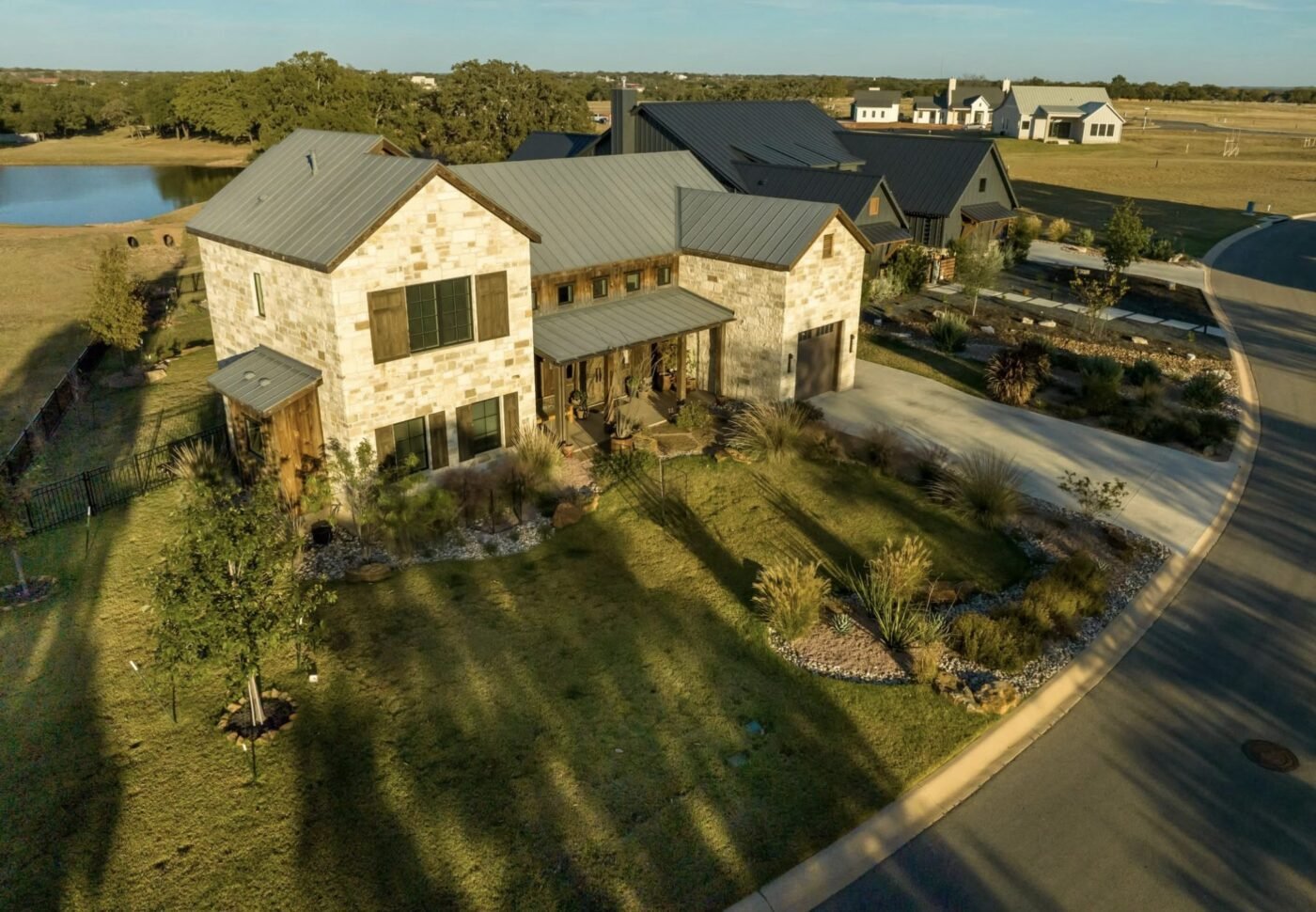 Two-story stone house with dark metal roof, porches, and landscaped front yard near a lake