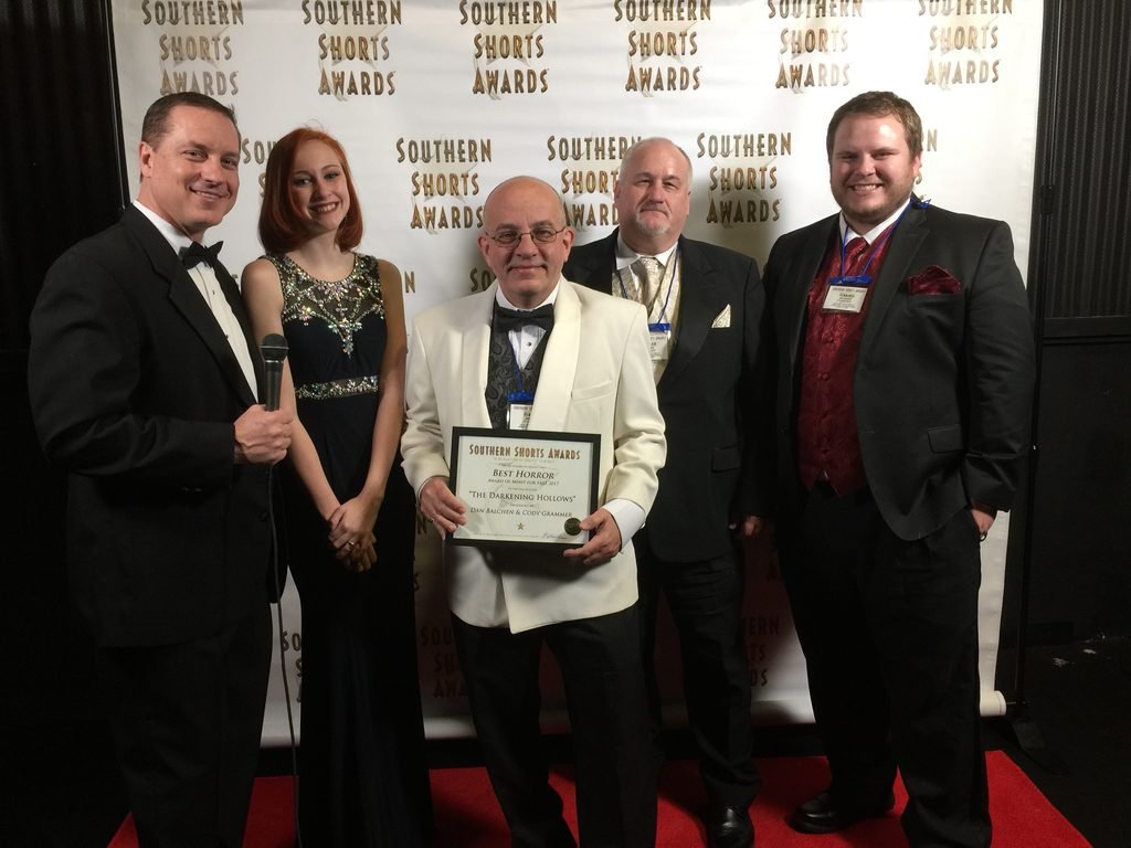 Five people in formal wear pose on a red carpet in front of a Southern Shorts Awards backdrop, with one person holding a plaque.