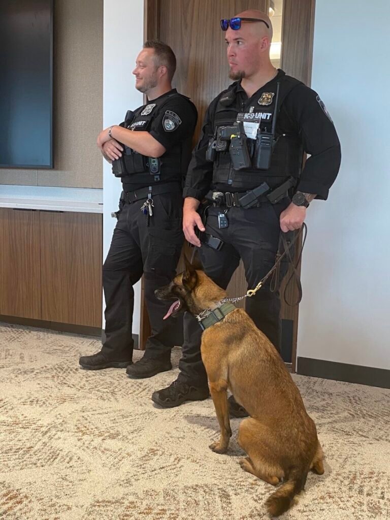 Two police officers stand with a seated brown working dog in a hallway