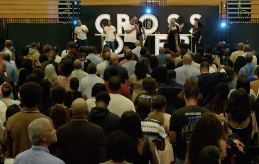 A crowd stands inside a church auditorium facing a stage with large illuminated letters spelling CrossOver