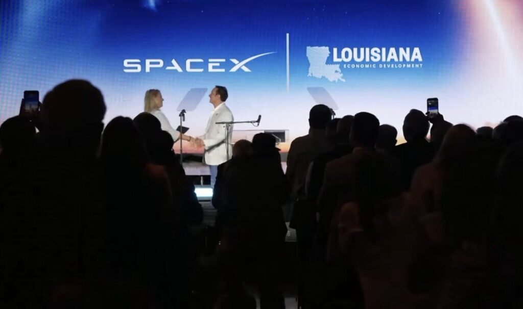 A crowd stands in silhouette at a stage where two people shake hands in front of SpaceX and Louisiana Economic Development logos