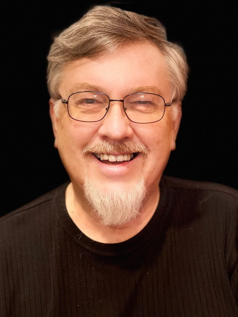 A smiling man with glasses and a goatee poses against a dark background.
