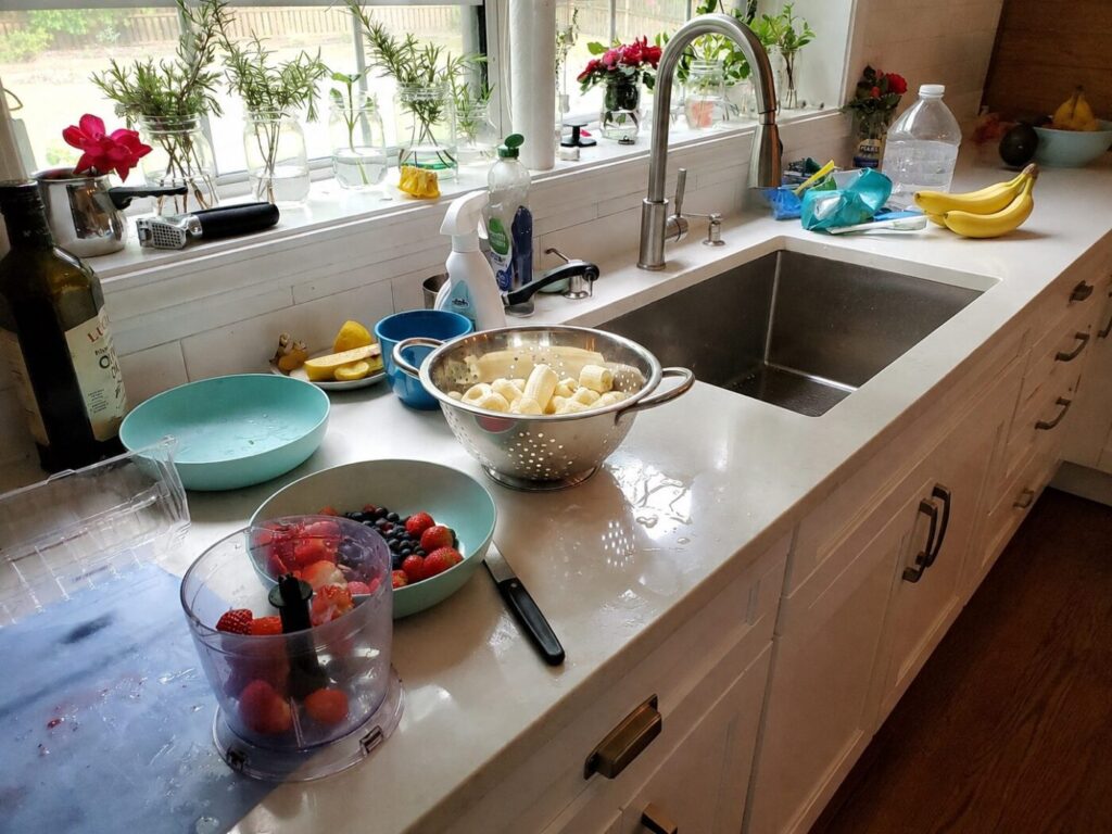 Kitchen sink with a tall faucet, dishes, fruit, and cleaning supplies on the counter