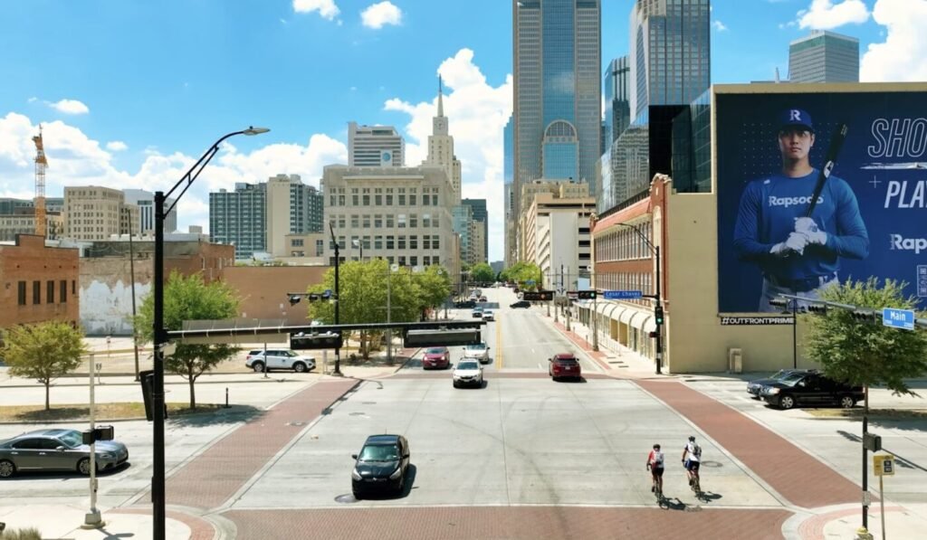 A downtown street runs toward a cluster of tall buildings, with a billboard on one side and cyclists crossing the intersection.