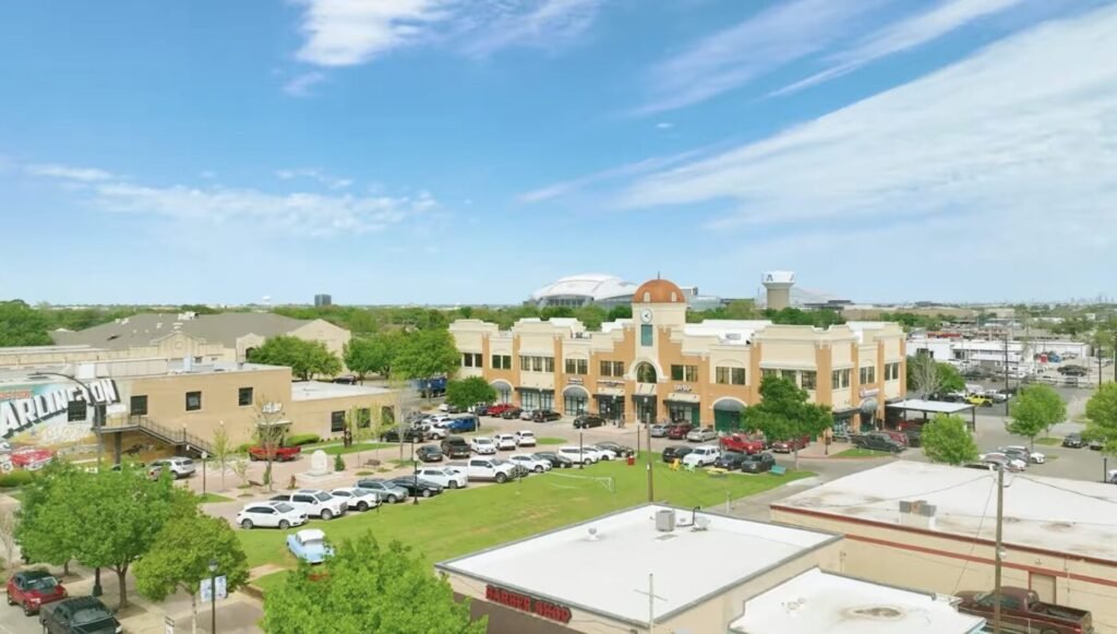 A shopping center and parking lot sit beside low-rise buildings under a blue sky.