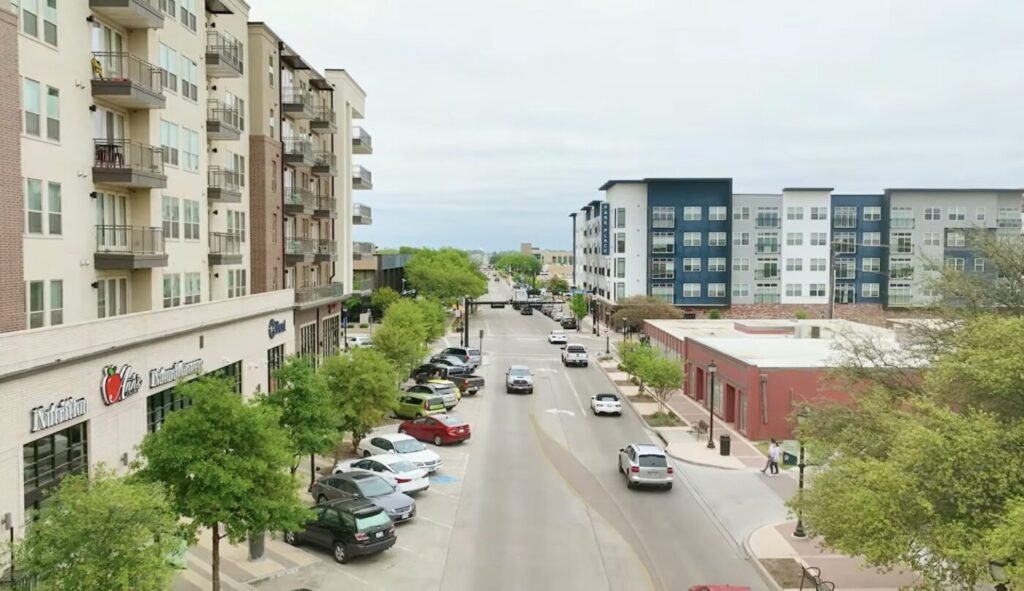 An urban street is bordered by apartment buildings, storefronts and parked cars.