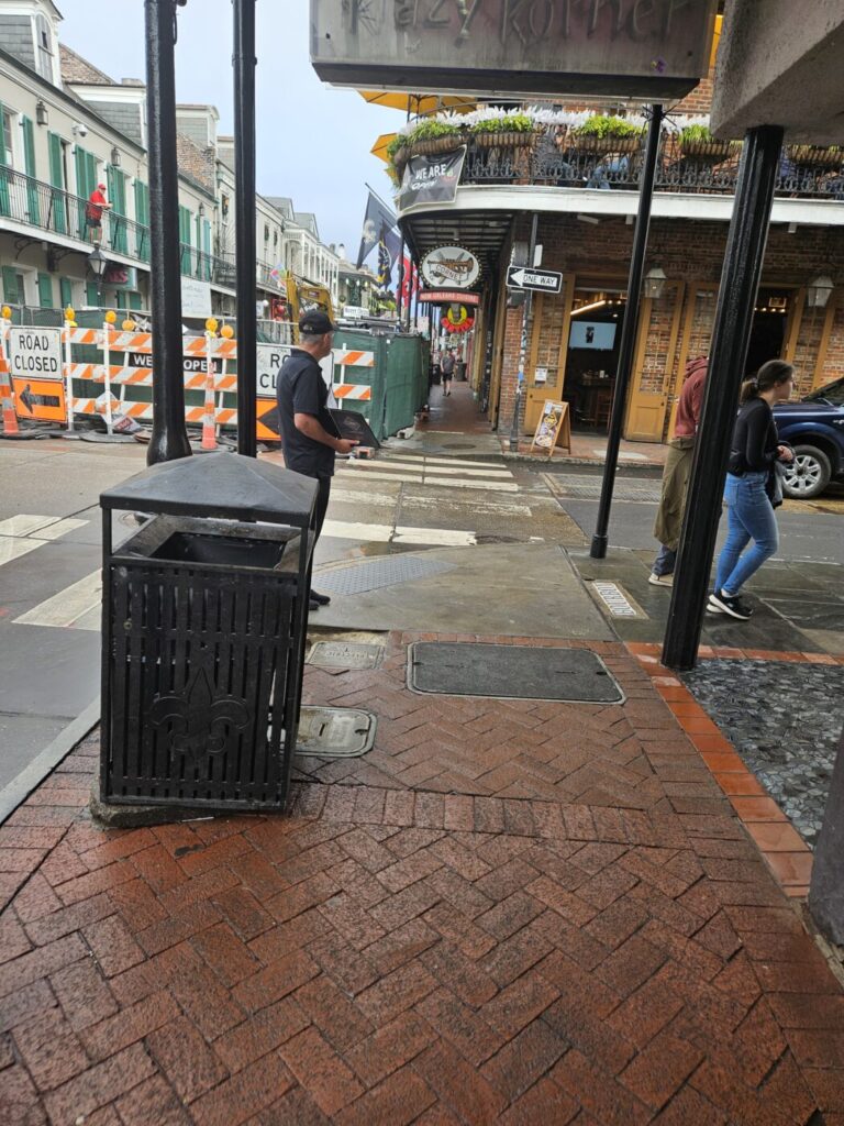 A wet brick sidewalk corner beside a closed street, with pedestrians, a trash can and construction barriers nearby.