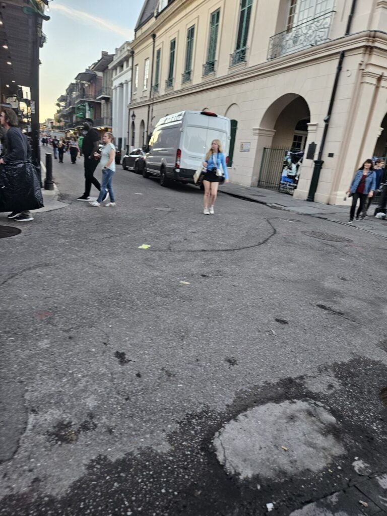 People walk along a street lined with historic buildings, parked vehicles and a white van.
