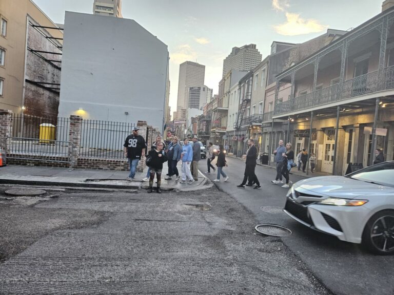People cross a worn city intersection between historic buildings and a passing car.