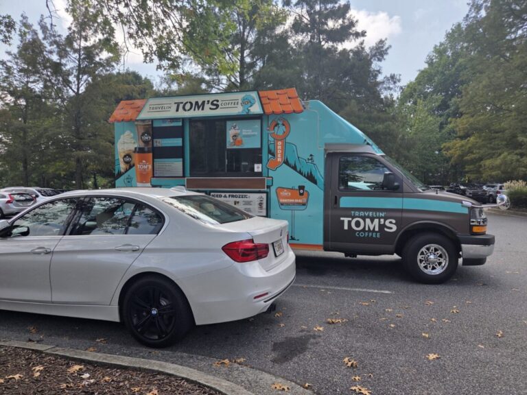 Turquoise Travelin’ Tom’s Coffee truck parked beside a silver sedan in a tree-lined lot.