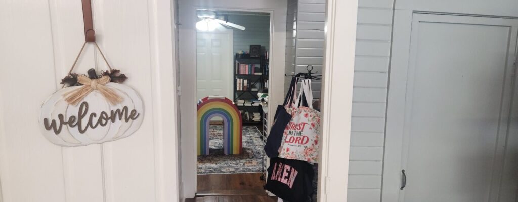 Welcome sign beside an open doorway with tote bags and a rainbow decoration inside