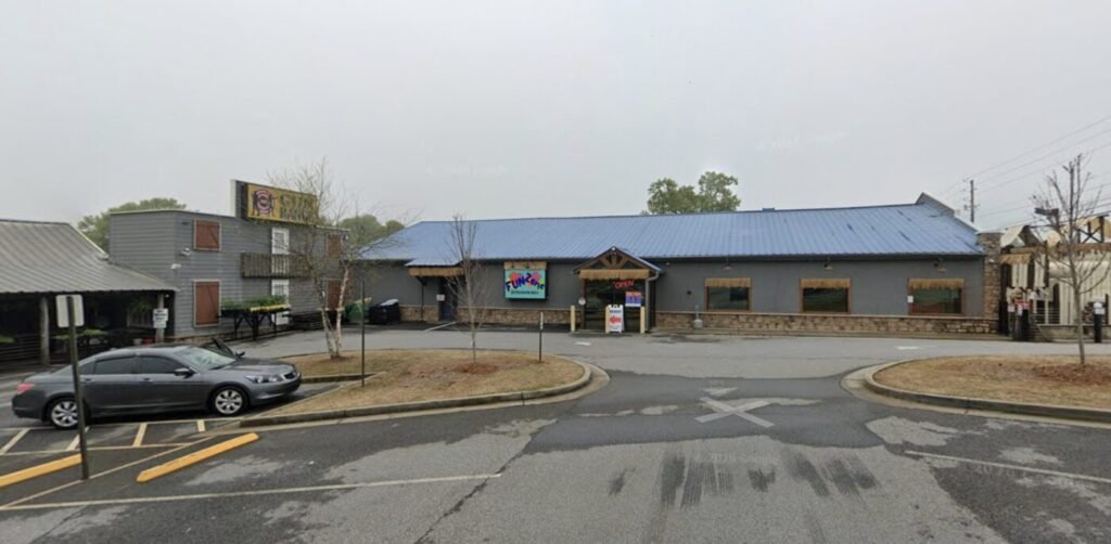 Wide view of a low commercial building with a blue roof and a parking lot in front