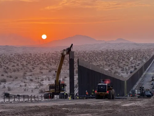 Bulldozers work beside a border wall at sunset in a desert landscape.