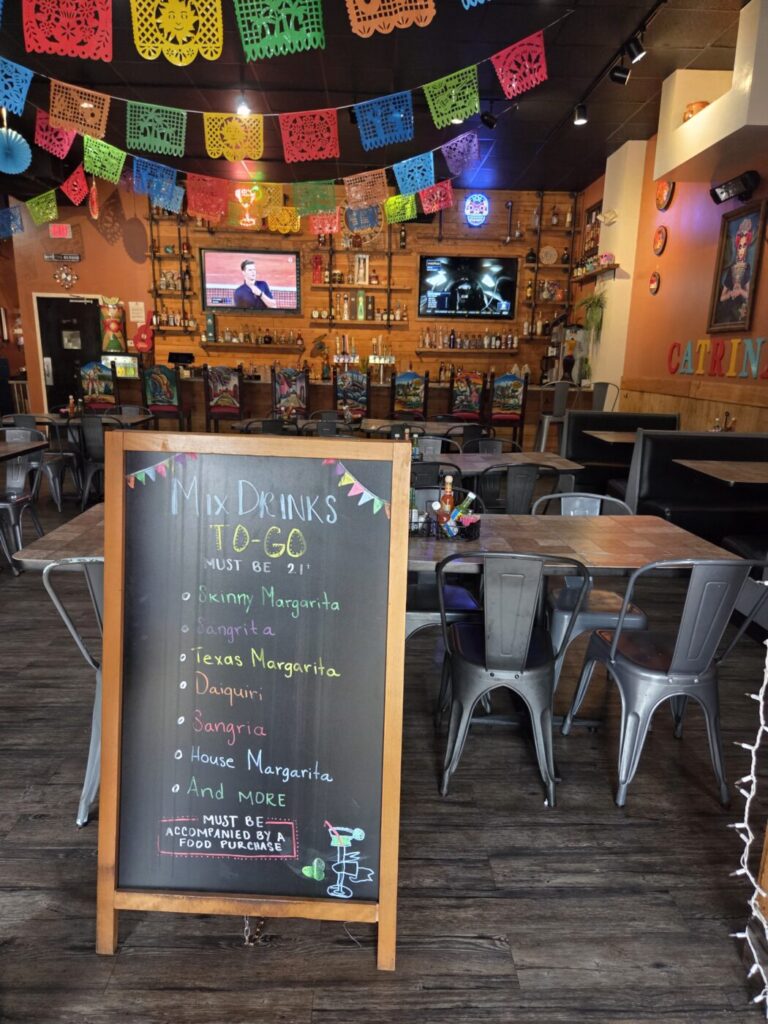 Colorful papel picado hangs over empty tables inside a Mexican restaurant, with a chalkboard announcing to-go drinks in the foreground.