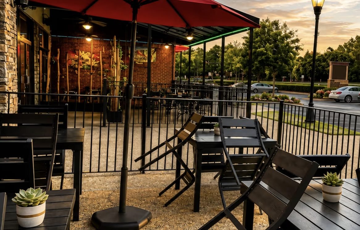 Outdoor patio seating with black tables and chairs under red umbrellas outside a brick restaurant at sunset
