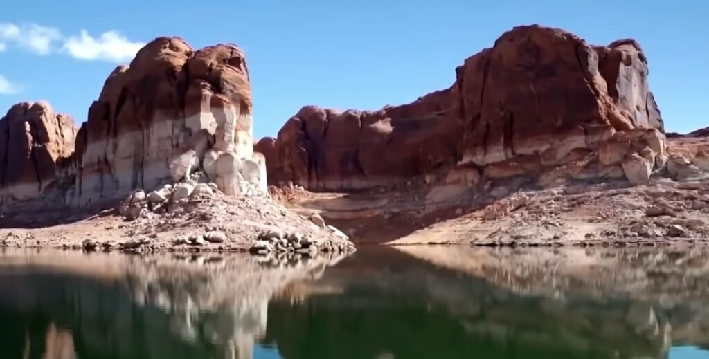 Red sandstone cliffs rise above calm reservoir water under a blue sky.