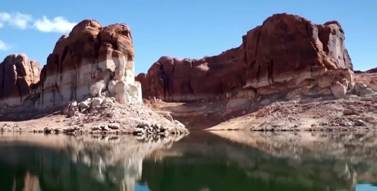 Red sandstone cliffs rise above calm reservoir water under a blue sky.